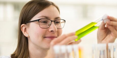 Michelle steht im Labor vor einem Ständer mit Reagenzgläsern und hält zwei mit grüner Flüssigkeit in der Hand.