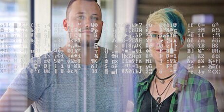 Erik und Jennifer stehen nebeneinander und blicken in die Kamera. Quasi vor ihren Gesichtern befindet sich eine Glasfläche mit IT-Symbolen.